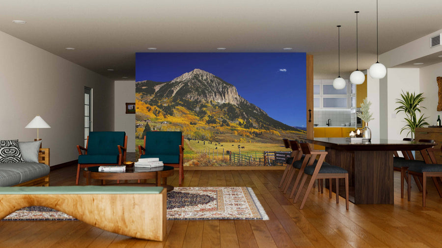 A modern, open living-dining space with wooden floors and mid-century furniture features the Crested Butte Mountain Over Ranch Wall Mural. Warm, natural-toned decor and globe pendant lights enhance the inviting atmosphere.