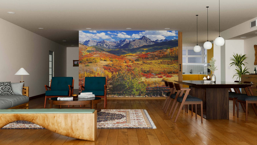A modern living-dining room with wood flooring, stylish furniture, pendant lights, and a large Dallas Divide Rolling Hills Wall Mural of colorful autumn mountains. Teal armchairs, a tan sofa, and a wooden dining table complete the inviting space.