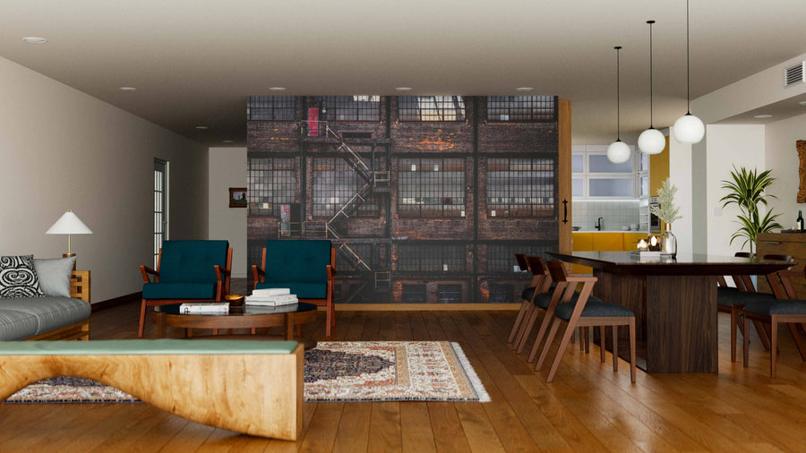 A modern living-dining room with wood floors, a gray sofa, and dark blue chairs features the District Wall Mural depicting an industrial building with windows and fire escapes, adding artistic flair to the space.