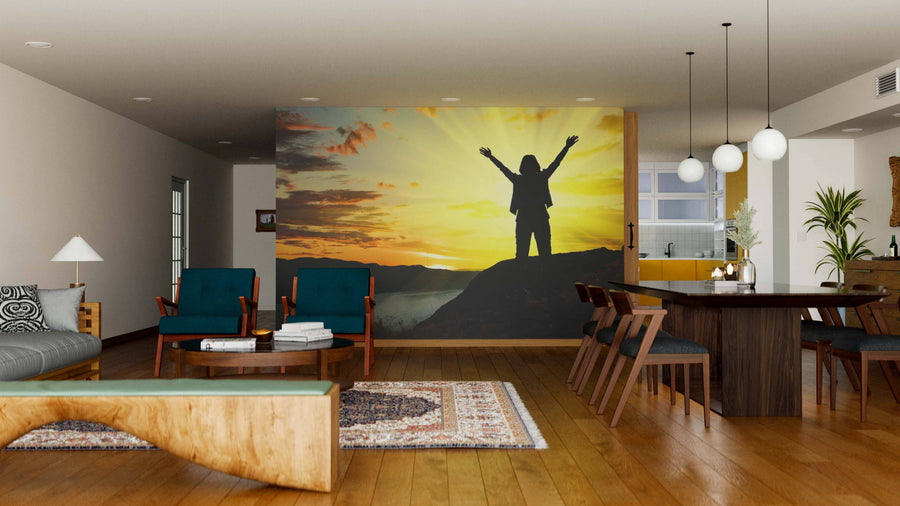 A modern living and dining area with wood flooring features the Dusk Wall Mural, showing a person with raised arms at sunset. Contemporary furniture, pendant lights, and houseplants add comfort with natural tones and textures.
