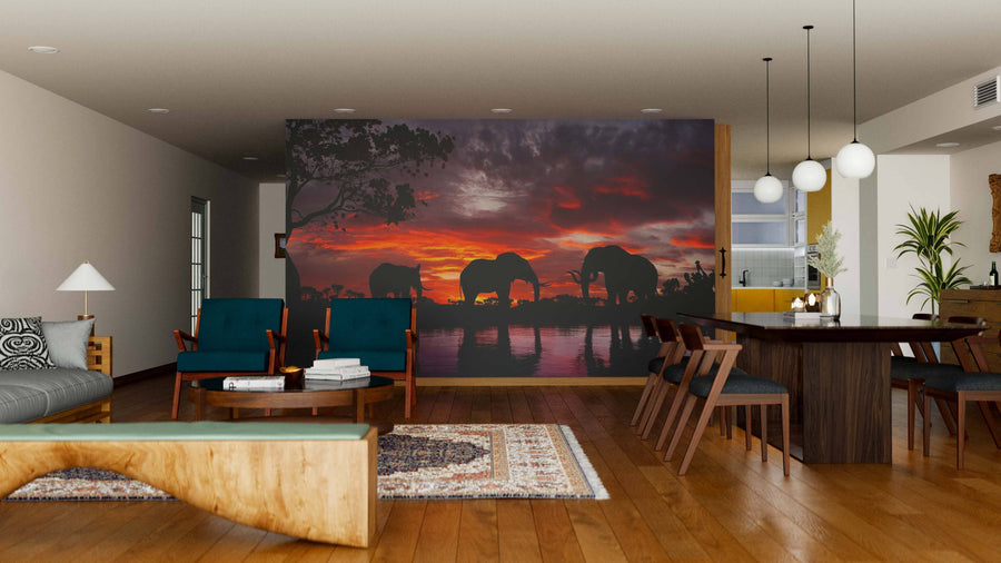 A modern living-dining room features wooden furniture, teal armchairs, and a rug. The Elephants At Sunset Wall Mural adds drama with silhouetted elephants against a fiery red and orange sunset, creating a striking focal point in the space.