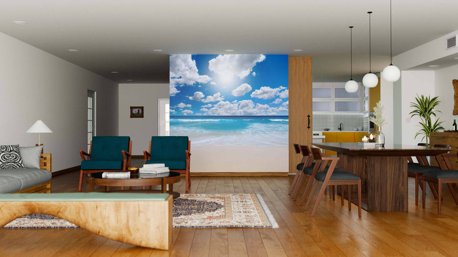 A modern open living-dining area with wood floors and furniture features the Gorgeous Beach Landscape Wall Mural, showing a blue sky and clouds. The space includes a gray sofa, blue armchairs, wooden table, hanging lights, and kitchen view.