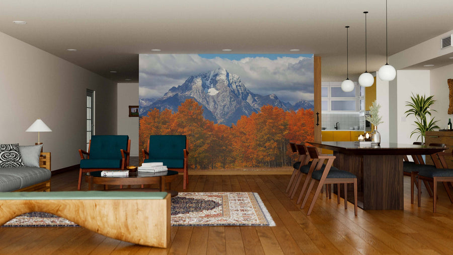 A modern open-plan living and dining room with wooden floors, mid-century furniture, and a Grand Teton Mountains Wall Mural depicting snow-capped peaks above autumn trees. Pendant lights hang over a dark dining table; a cozy seating area sits to the left.