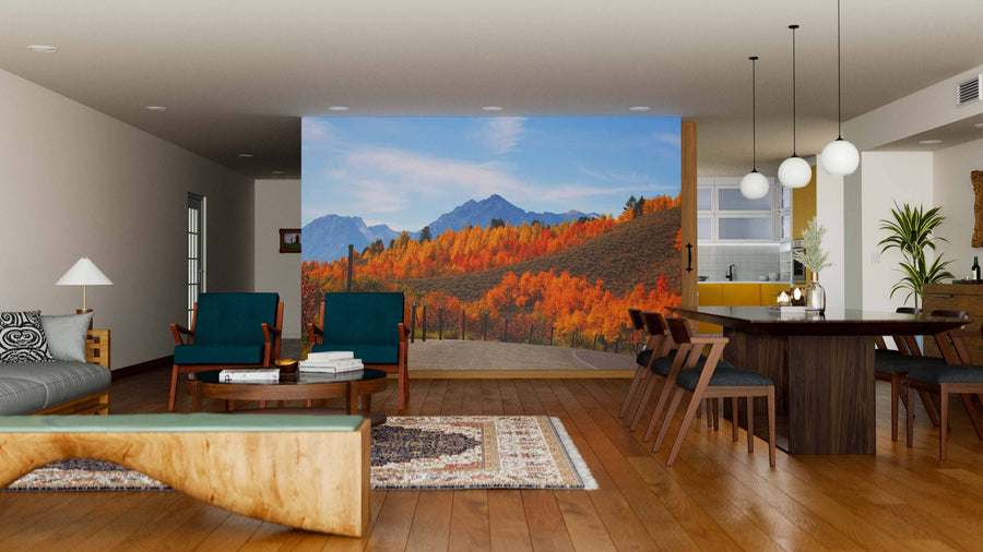 A modern living and dining room with wood floors, green chairs, a wooden table set, and the Grand Teton Mountain Range Wall Mural showcasing an autumn mountain landscape with a blue sky and vibrant red, orange, and green trees.