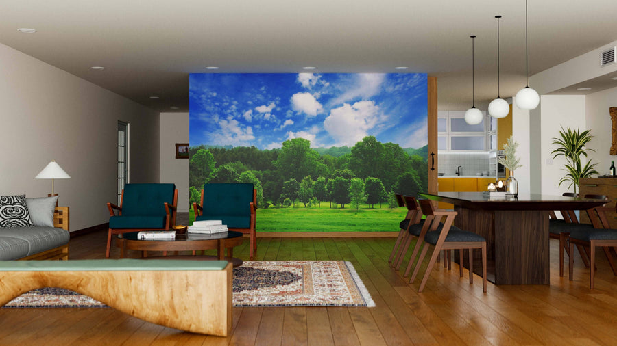 A modern open living-dining room features wood floors, green and wood furniture, the Green Forest Open Field Wall Mural on one wall, pendant lights above a dark dining table, and a kitchen with white cabinets in the background.