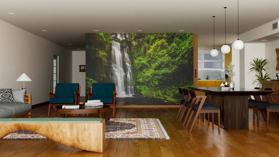 A modern living and dining area with wood floors, mid-century furniture, and a Maclean Falls Wall Mural depicting a lush green forest and waterfall. The space includes blue chairs, a wooden table, floor rug, and hanging globe lights.