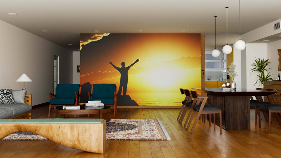 A modern living and dining room with wooden floors, teal chairs, hanging lights, and the Mean Greeting Sun Wall Mural depicting a person’s silhouette on rocks against a vivid yellow-orange sunset.