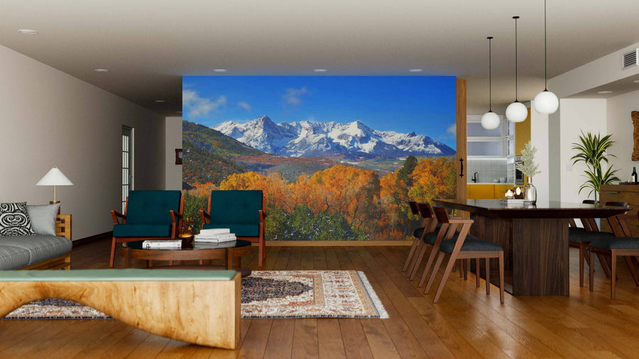 A modern living-dining room features wooden floors, green armchairs, a large dining table, and globe pendant lights. The Mount Sneffels Panoramic View Wall Mural displays snow-capped peaks above autumn trees as a stunning nature-inspired focal point.