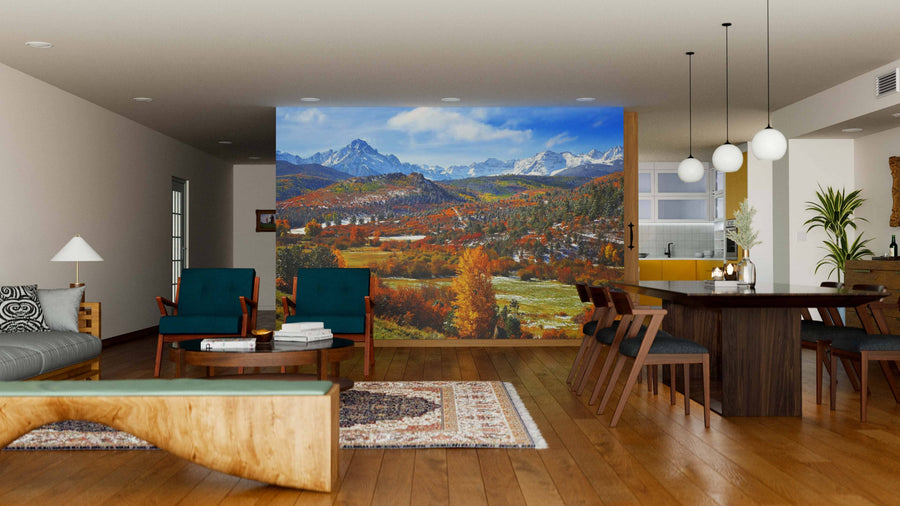 A modern open-concept living and dining room with wood floors, a rustic bench, green and brown furniture, round pendant lights above the table, and a large Mount Sneffels in The Fall Wall Mural of a colorful autumn landscape.