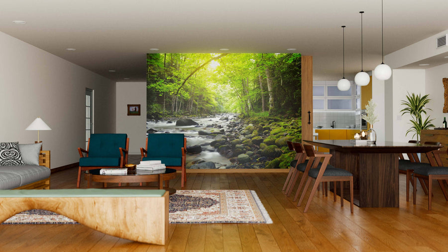 A modern living-dining space with wooden floors, blue and wood furniture, pendant lighting, and a large Mountain River in the Wood Wall Mural depicting a sunlit forest and stream for a serene, inviting ambiance.
