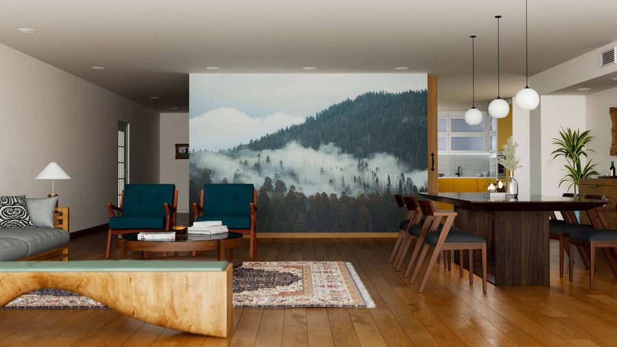 Modern living-dining room with wood floors, teal chairs, patterned rugs, and a large Mountain with Fog Wall Mural. Pendant lights hang above a dark dining table; the kitchen is visible behind a glass wall in this earthy space.