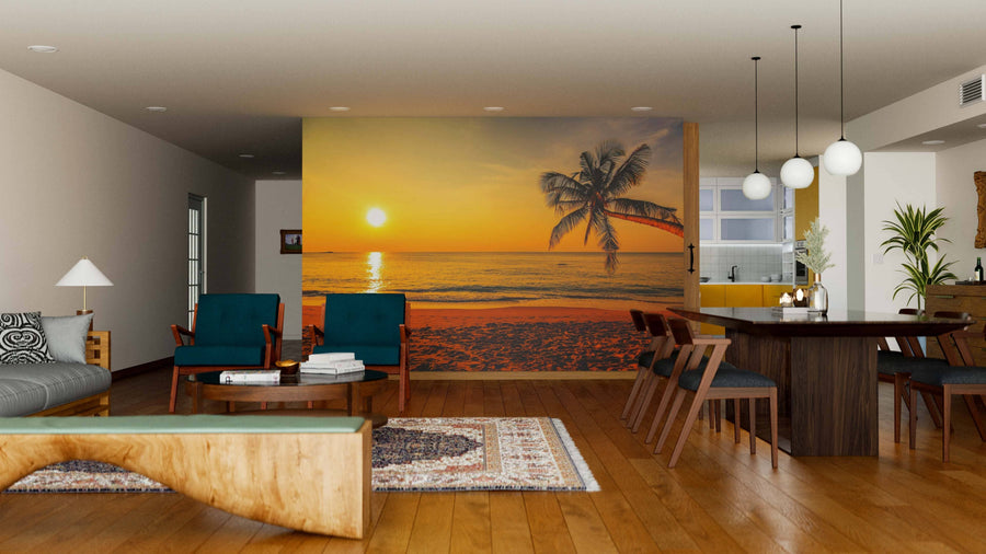 A modern open-plan living and dining room with wooden floors and mid-century furniture is brightened by the Nice Sunset Wall Mural, featuring a vibrant beach sunset with a palm tree. Sunlight filters through pendant lights above the dining table.