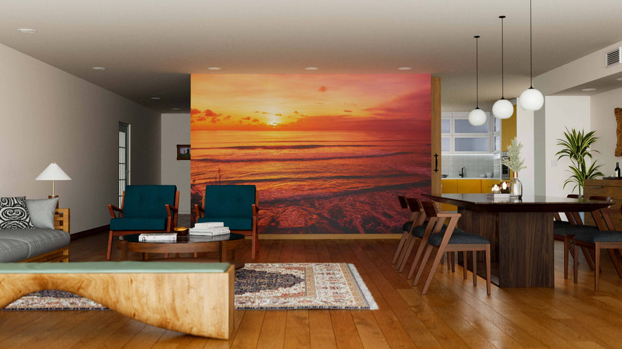 A modern living-dining area with wooden floors, teal armchairs, and an Ocean Waves Wall Mural depicting an orange sunset over the ocean. Cozy lighting, a rug, and round pendant lights add warmth and style.