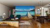 A modern living-dining room with wooden floors, teal armchairs, and pendant lights is highlighted by the Old Wooden Pier Wall Mural—a peel and stick mural of a pier extending into turquoise sea beneath a blue sky with clouds.