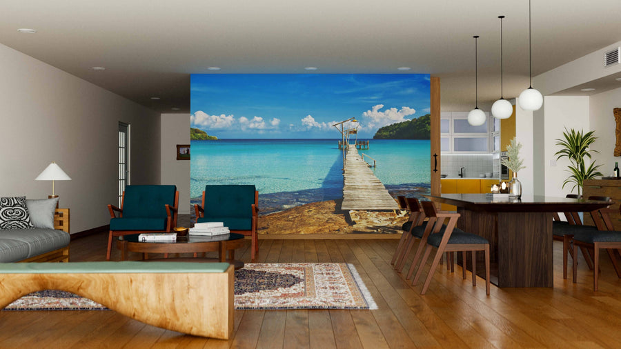 A modern living-dining room with wooden floors, teal armchairs, and pendant lights is highlighted by the Old Wooden Pier Wall Mural—a peel and stick mural of a pier extending into turquoise sea beneath a blue sky with clouds.