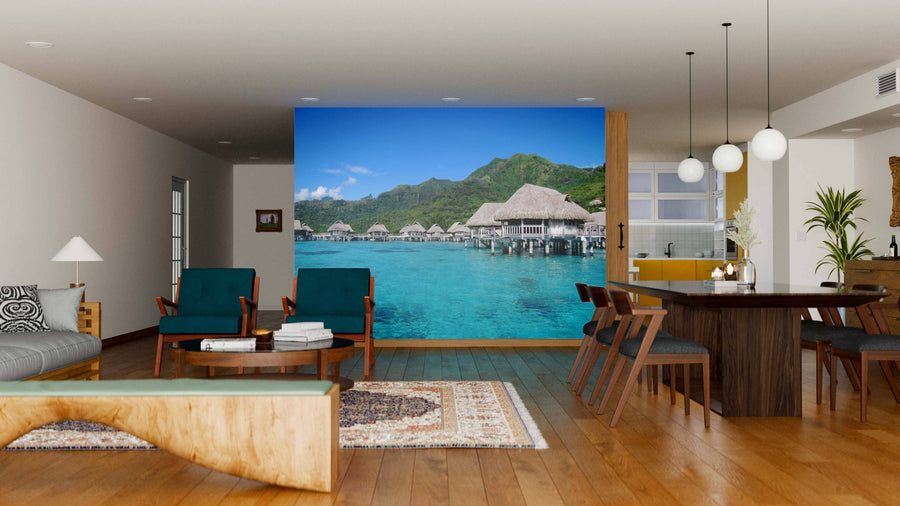 A modern living and dining room features wood floors, mid-century furniture, and the Overwater Bungalow Wall Mural depicting turquoise water. Neutral tones and pendant lights create a warm, tropical-inspired atmosphere.