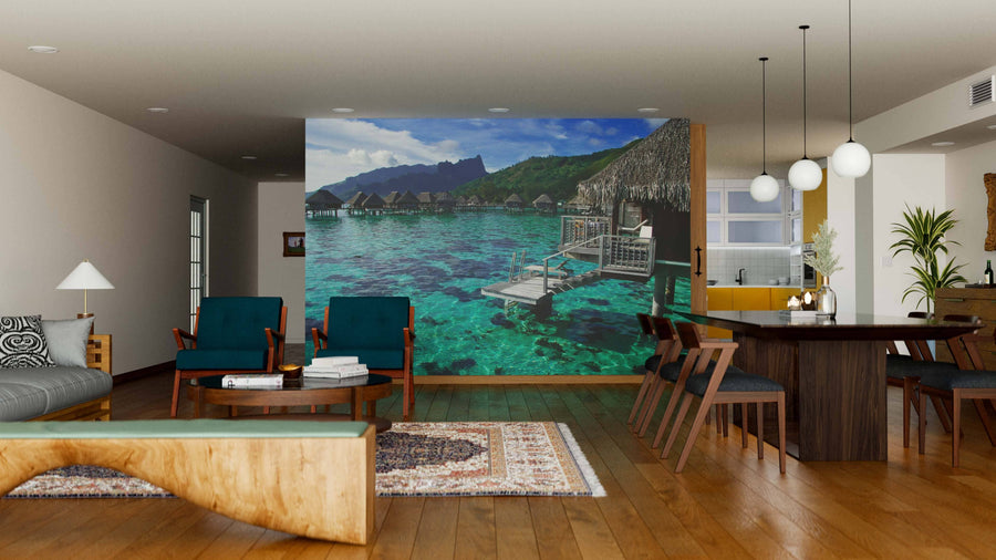 A modern living and dining room with wood floors, a rug, chic seating, and a unique coffee table is elevated by the Overwater Bungalows in French Polynesia Wall Mural, showcasing turquoise water, bungalows, and mountains for a tropical touch.