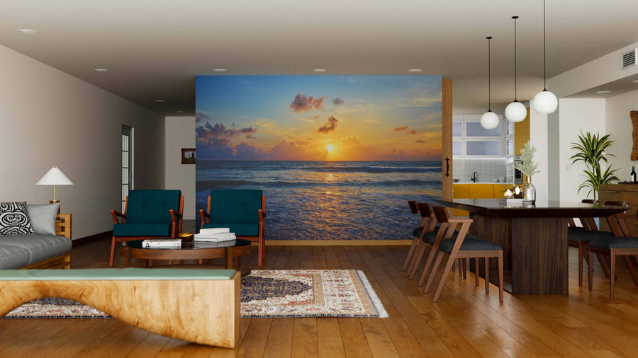 A modern open-plan living and dining room with wooden floors, teal armchairs, a dark wood table, and pendant lights features the Phuket Beach Wall Mural—a peel and stick mural of a serene ocean sunset that adds warmth and relaxation to the space.