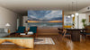 A modern open living-dining room with wood flooring, teal armchairs, and a dark wood dining table features a dramatic sunrise view created by the Picturesque Morning Scene Wall Mural. Pendant lights are suspended above the table.