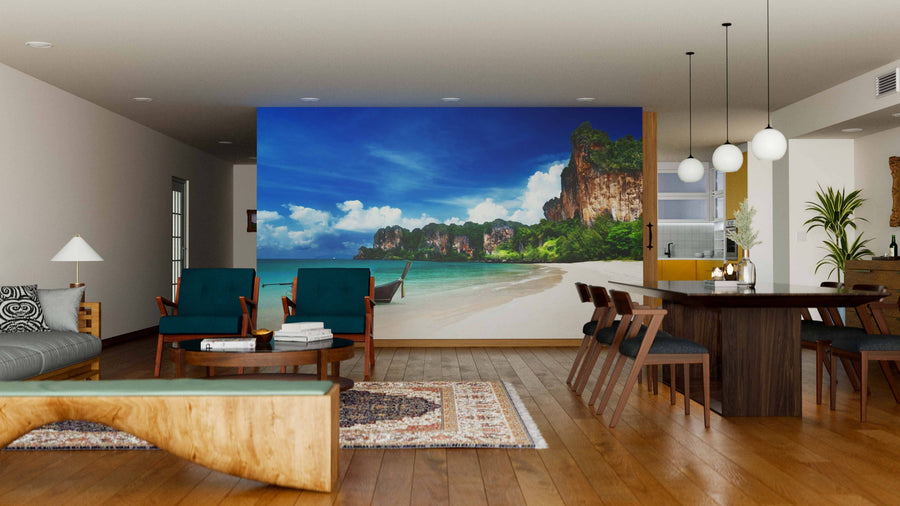 A modern living-dining room with wood floors and teal chairs features the “Railay Beach in Krabi Thailand Wall Mural,” depicting a tropical beach. Globe lights, a sofa, rugs, and natural decor complete this inviting space.