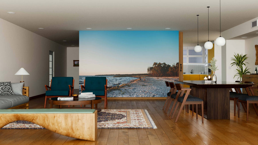 A modern, open-plan living and dining area features wooden floors, blue armchairs, a gray sofa, rustic bench, dining table with chairs, pendant lights, kitchen in the background, and a large Sandy Sea Coast Wall Mural.