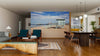A modern living-dining room features wooden floors, mid-century furniture, and a Santa Monica Beach and Pier Wall Mural, creating an open coastal vibe. Pendant lights hang above the table and a rug accents the wood décor.