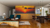 Modern living and dining room with wooden floors and furniture, featuring the Sea Sunset Deep Red Wall Mural depicting a vivid orange sunset over the ocean. The space has teal armchairs, a bench, rugs, and globe pendant lights above the dining table.