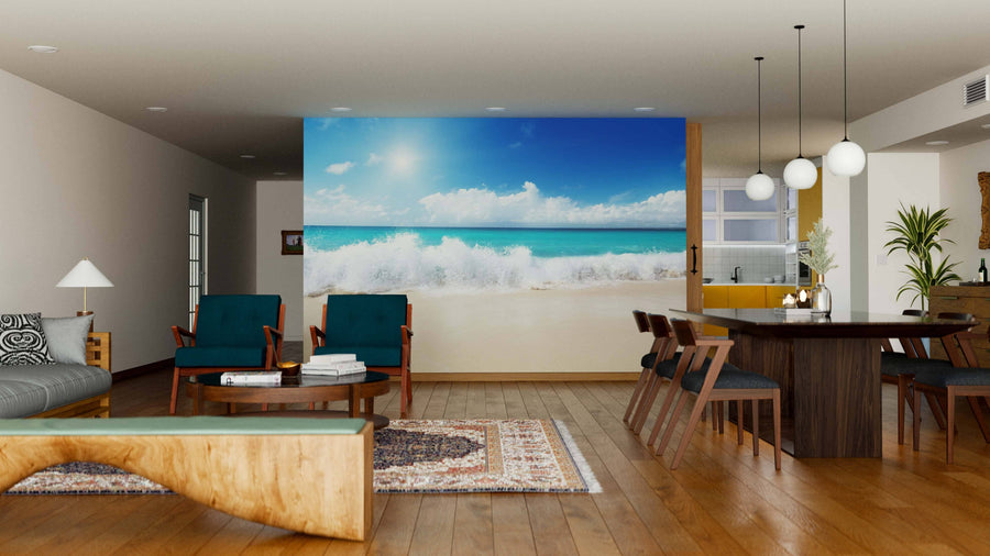 A modern, open-plan living and dining area with wooden floors, teal armchairs, a rug, and the Sea and Sand Wall Mural depicting a sunny beach with waves for a seaside feel. Spherical pendant lights hang above the dining table.