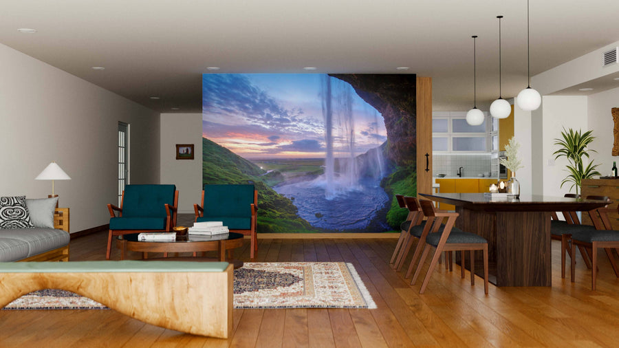 A modern living-dining room with mid-century furniture and wooden floors features the Seljalandfoss Waterfall Wall Mural on an accent wall, bringing natural beauty and a sunset landscape into the open, neutral-toned space with contemporary lighting.