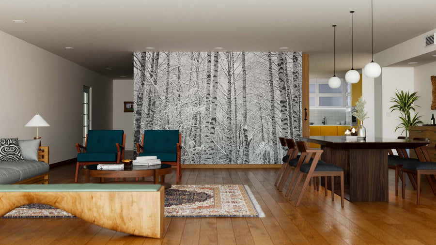 A modern open-plan living and dining area with wood floors, teal armchairs, a large table, and a decorative rug is accented by the Snow on the Branches Wall Mural, bringing a striking black-and-white birch tree design to the cozy interior.