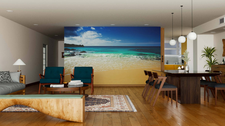 A modern living-dining room with wood floors, teal armchairs, and a dining table under globe lights is accented by the Stranded Beach Wall Mural, which adds a striking blue sky and ocean focal point to the open, airy space.
