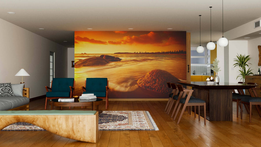 A modern living-dining room features wooden floors, mid-century furniture, and a Sun Tinted Beach Wall Mural of an orange sunset over ocean rocks. Pendant lights hang above a dark dining table, while cozy rugs add warmth to the inviting space.