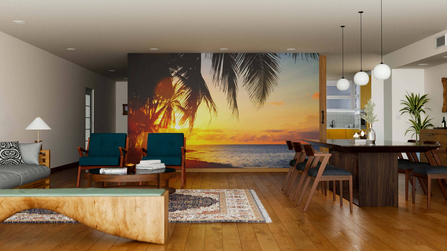 A modern living-dining room with wooden floors, blue armchairs, and pendant lights features the "Sunrise Over the Tropical Beach Wall Mural" on the back wall, displaying palm trees and a vivid orange sky above the sea at sunrise.