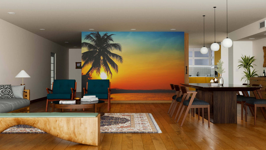 Modern living-dining room with wooden floors, teal chairs, a rug, and a large Sunrise in the Tropics Wall Mural featuring a palm tree over the ocean; pendant lights hang above the dark dining table with the kitchen visible behind.