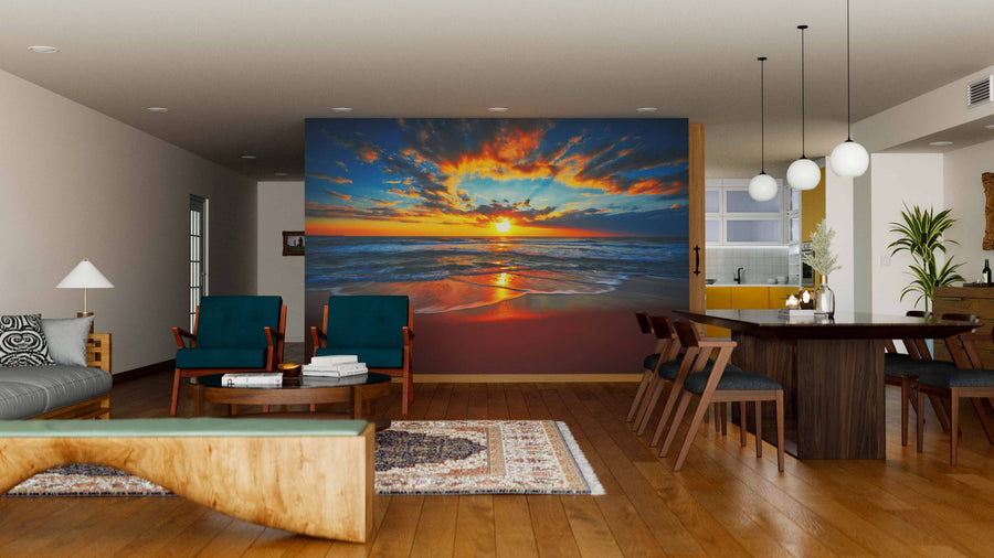 A modern living-dining area with wooden floors features teal armchairs, a wooden table, and the Sunrise on the Beach Wall Mural, depicting a vibrant ocean sunset. Pendant lights illuminate the dining spot, with the kitchen in view.