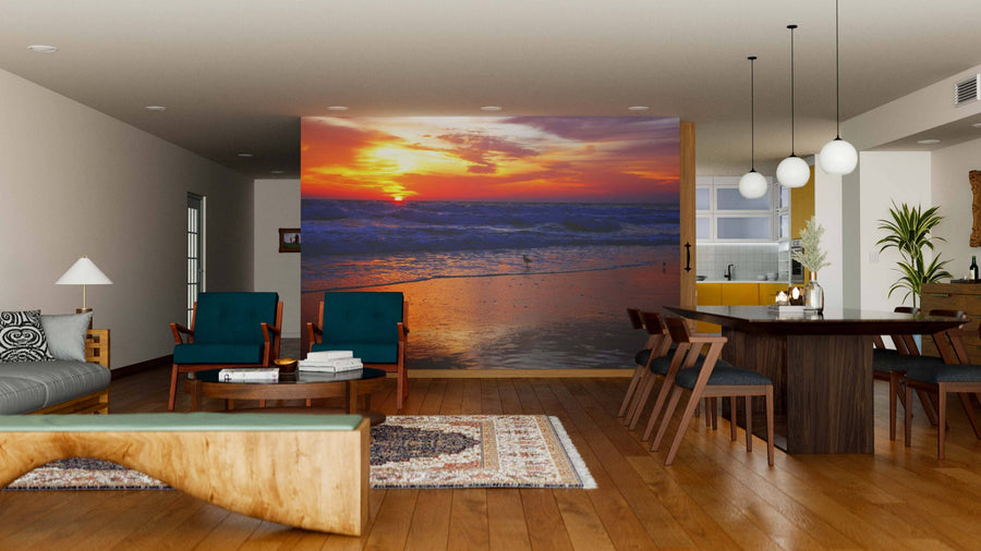 A modern living-dining room features wooden floors, stylish furniture, pendant lights, and a wall-sized Sunrise on the Surf Wall Mural, showcasing a vibrant ocean sunrise that adds warmth and color to the open-concept space.