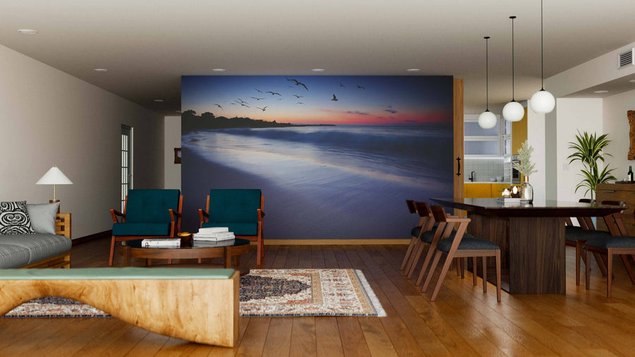 A modern living-dining room with wooden floors, teal chairs, a wooden bench, rugs, and the Sunset Beauty Wall Mural showing a beach sunset with birds. Pendant lights hang above a dark dining table, with a sideboard in the background.