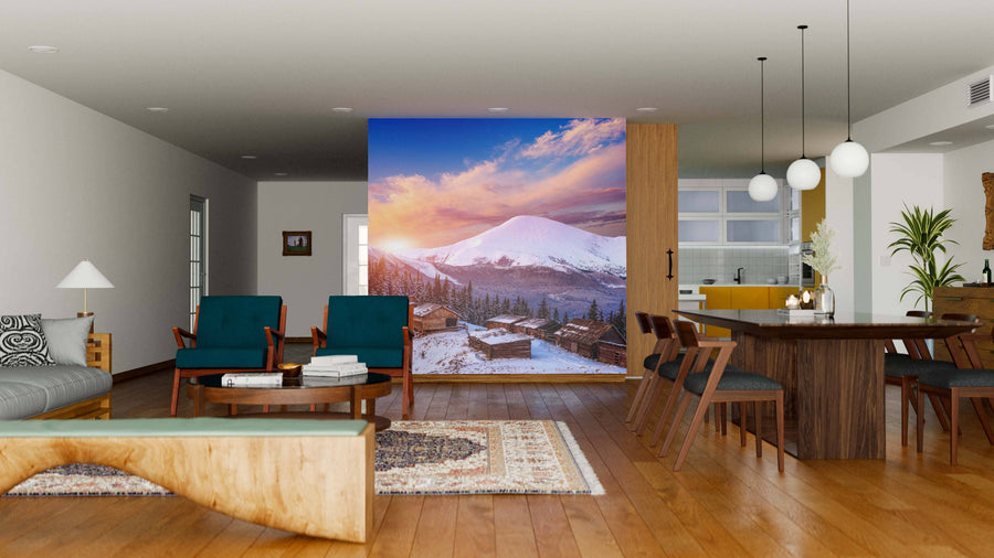 A modern living-dining area with wood floors, teal armchairs, and pendant lights features the Sunset Over Mountain Cottage Wall Mural, depicting a snow-capped mountain at sunset above a village and adding color to the neutral space.