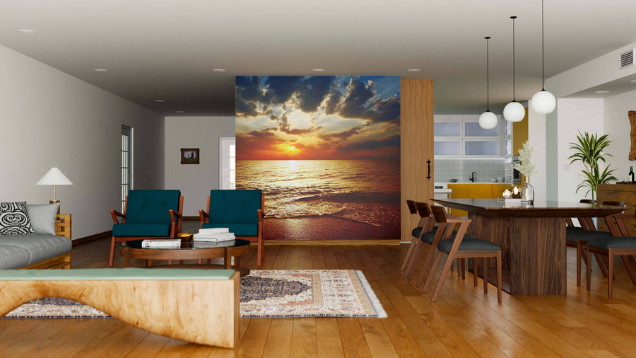A modern open-plan living and dining room with wood floors, teal armchairs, and a kitchen features the Sunset Over Sea Wall Mural, whose vibrant ocean sunset with clouds adds warmth and color to the decor.