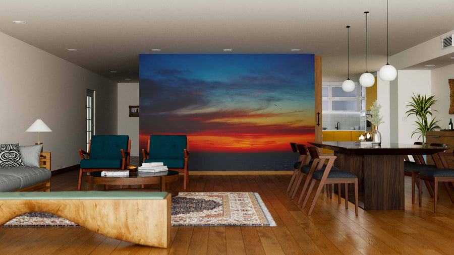 A modern living and dining area with wood floors, teal armchairs, a large dining table, and pendant lights features the Sunset Over the Ocean Wall Mural, bringing vibrant color and warmth to this open, stylish space.