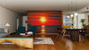 A modern living-dining room with wood floors, blue and brown furniture, pendant lights, and the Sunset at the Beautiful Beach Wall Mural featuring a vivid red-orange ocean sunset creates a dramatic focal point in the space.