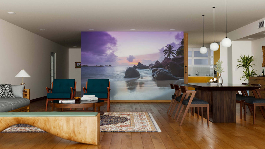 A modern living-dining room with wooden floors, teal armchairs, a unique bench, and dining table is enhanced by the “Sunset on Seychelles Beach Wall Mural,” featuring a tranquil rocky beach and purple sunset skies with palm trees.