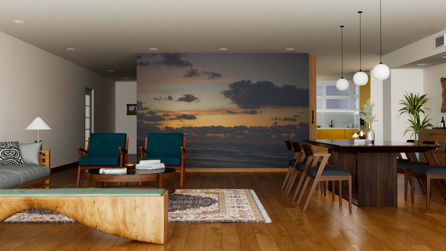 A modern open-plan living and dining room features wood floors, teal armchairs, and a long table, with the Sunset on the Battic Wall Mural—depicting a sunset over the ocean with clouds—creating a stunning backdrop behind the seating area.