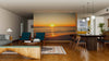 A modern living and dining area features wooden floors, stylish furniture, and cozy decor. The “Sunset on the Shore Wall Mural” fills the room with vibrant ocean sunset hues, complemented by pendant lights and warm-toned rugs for an inviting feel.