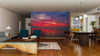 Modern living-dining room with wooden floors, teal and wood furniture, and a Sunset over Bay Wall Mural depicting a vivid red and purple seascape. Pendant lights brighten the dining area; the cozy seating includes a coffee table and rug.