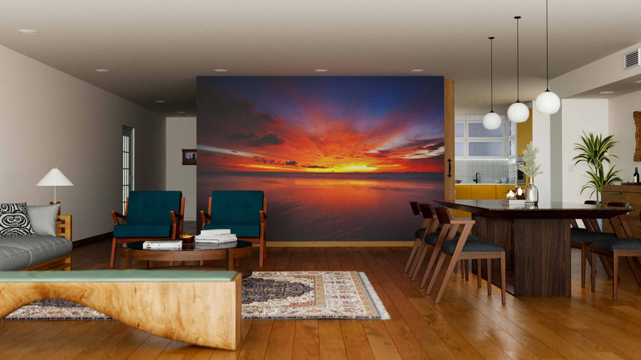 A modern living and dining room with wooden floors and mid-century furniture features the Tropical Sea Sunset Wall Mural, showcasing vivid orange, red, and purple clouds over the ocean as a striking focal point in the open space.