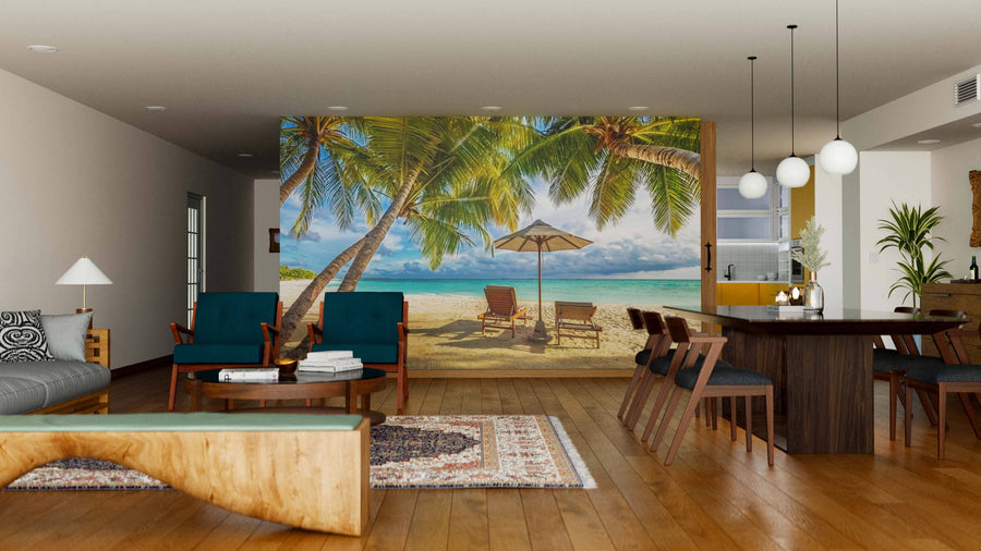 Modern living-dining room with wooden floors, dark teal furniture, and a large Tropical Sunset Scenery Wall Mural featuring palm trees, lounge chairs, sand, and ocean. Pendant lights hang above a wooden dining table with mid-century chairs.
