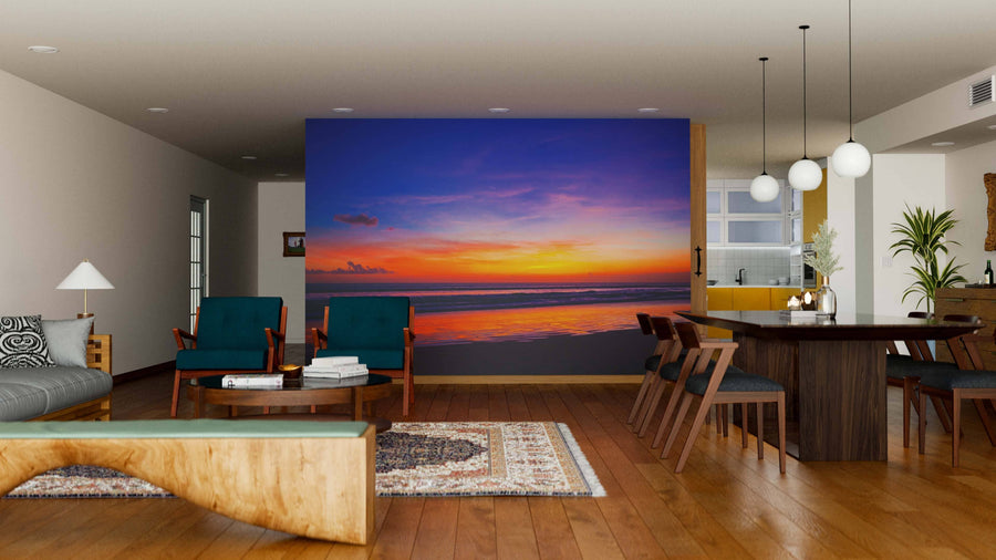 A modern living-dining room with wooden floors, teal chairs, a rug, and a sculpted coffee table features a vivid “Tropical Sunset on the Beach Wall Mural” in orange, purple, and blue tones above calm water on the back wall.