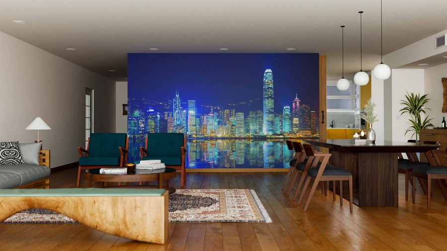 A modern living and dining space with wooden floors and furniture features the Victoria Harbor Wall Mural, displaying vibrant nighttime skyscrapers reflected on water. Warm hanging lights and rugs complete this inviting, stylish room.