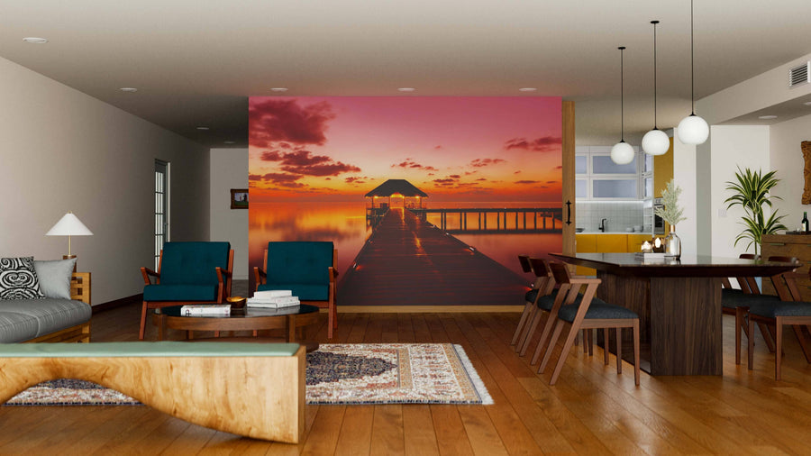 A modern living-dining area with wooden floors and stylish furniture features the "Water Cafe at Sunset Maldives Wall Mural," adding warmth. Pendant lights hang above the dining table, with a bookshelf and kitchen in the backdrop.
